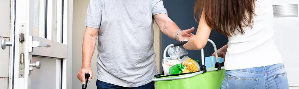 Aide à domicile remettant un panier de courses à une personne qui utilise une canne