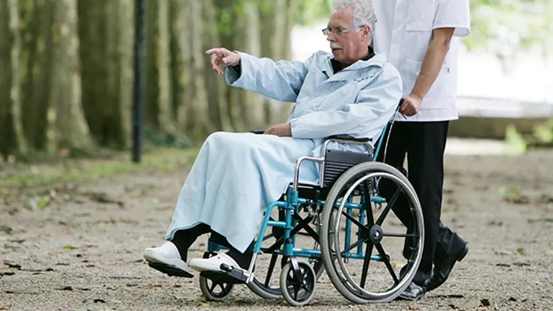 Personne âgée en fauteuil roulant accompagnée par un auxiliaire de vie sur une allée arborée